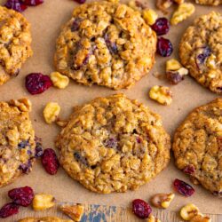 Oatmeal Cranberry Walnut Cookies laid out on brown parchment paper, with extra walnut pieces and dried cranberries