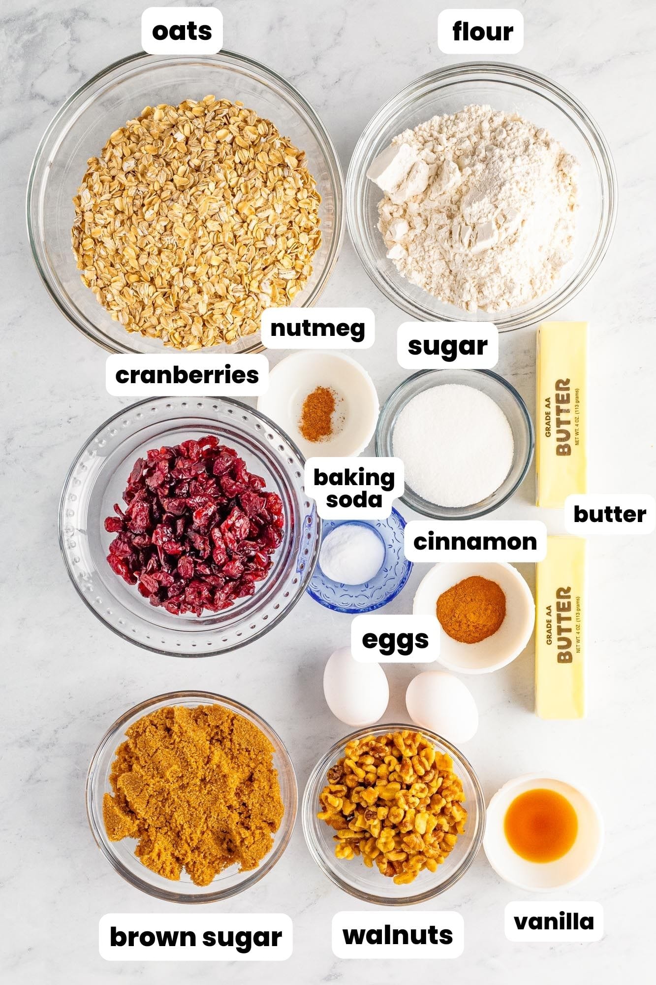 Ingredients needed to make oatmeal cranberry walnut cookies, including old-fashioned oats, all purpose flour, dried cranberries, plain sugar, brown sugar, walnut pieces, eggs, vanilla, butter, baking soda, salt, and some spices.