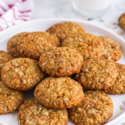 anzac cookies on a round white plate. In the background is a glass of milk.