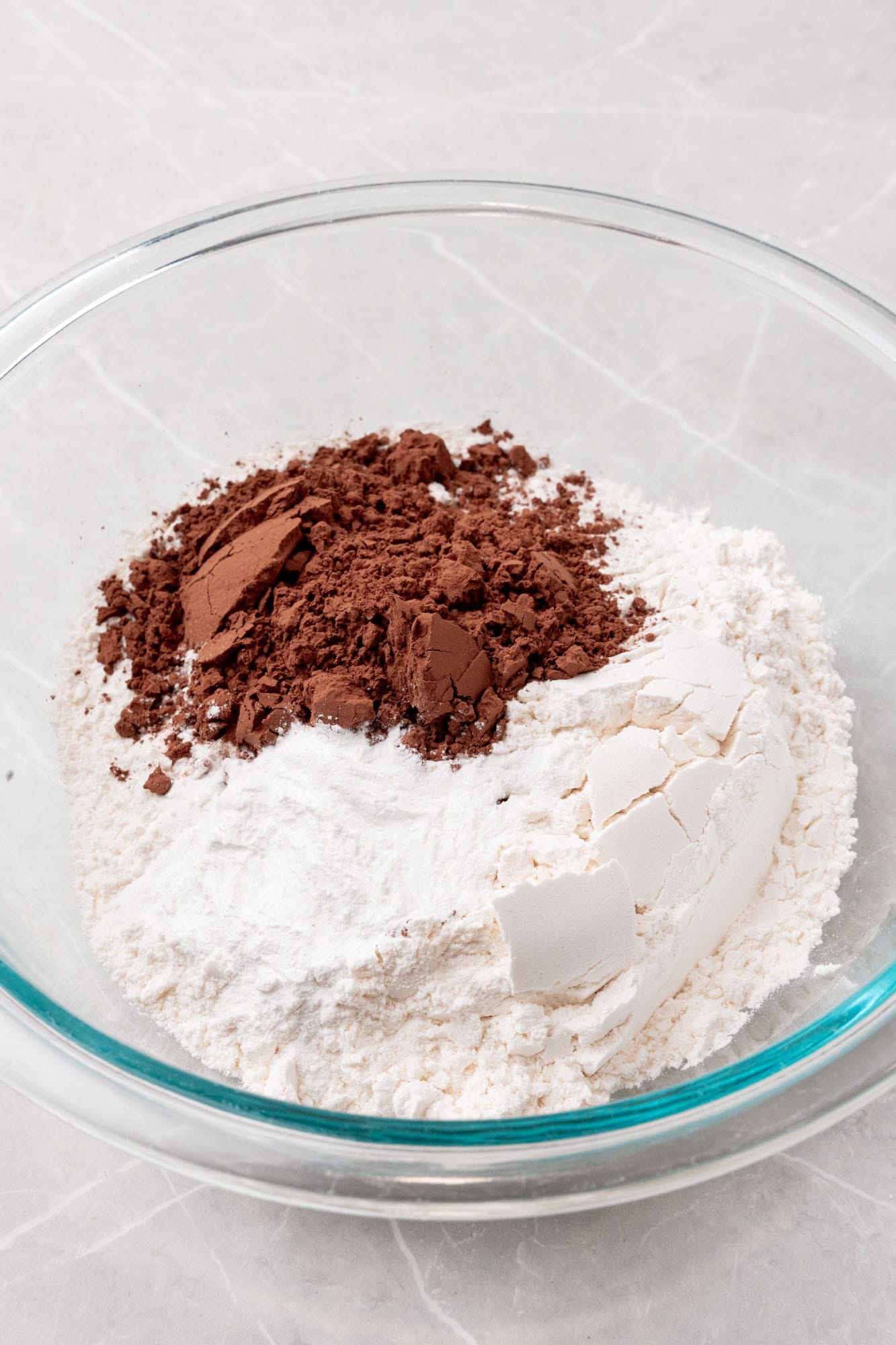 flour and cocoa powder in a glass mixing bowl on a marble counter.