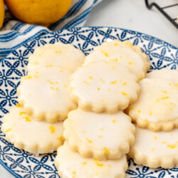 fluted circular lemon shortbread cookies on a blue and white plate next to fresh lemons.