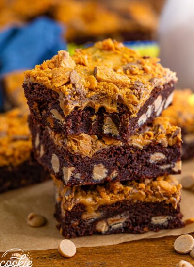 a stack of three brownies with peanut butter chips, topped with peanut butter and crushed butterfinger candies. The top brownie has a bite taken from the front corner.