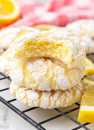 a stack of three lemon crinkle cookies on a wire rack. the top cookie has a bite taken to show the yellow, soft center.