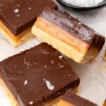 four squares of millionaire shortbread, showing three layers of cookie, caramel, and chocolate. in the background is a small black bowl of salt.