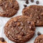 Chocolate fudge cookies with crinkly tops on a piece of parchment paper. Chocolate chips are arranged around the cookies.