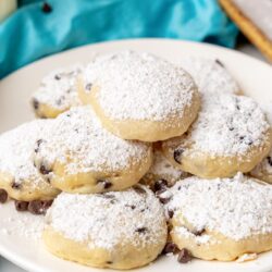 a white plate filled with cheesecake cookies with chocolate chips. The cookies are dusted with powdered sugar.
