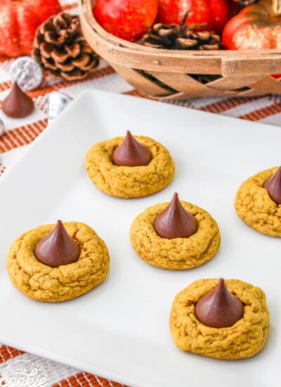 Freshly baked pumpkin spice blossom cookies on a white plate, each topped with a chocolate kiss, set on an autumn-themed table with pumpkins and pinecones in the background.