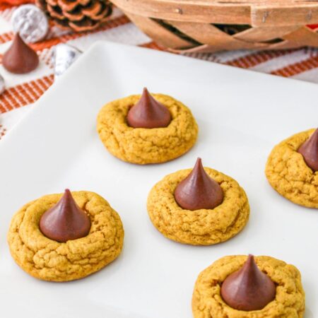Freshly baked pumpkin spice blossom cookies on a white plate, each topped with a chocolate kiss, set on an autumn-themed table with pumpkins and pinecones in the background.