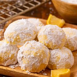 Lemon snowball cookies coated in powdered sugar stacked on a wooden board with fresh lemon wedges and a bowl of powdered sugar in the background