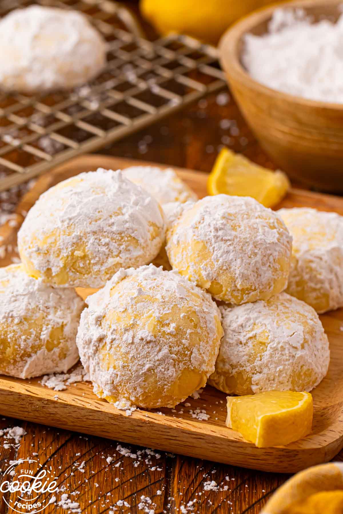 Lemon snowball cookies coated in powdered sugar stacked on a wooden board with fresh lemon wedges and a bowl of powdered sugar in the background