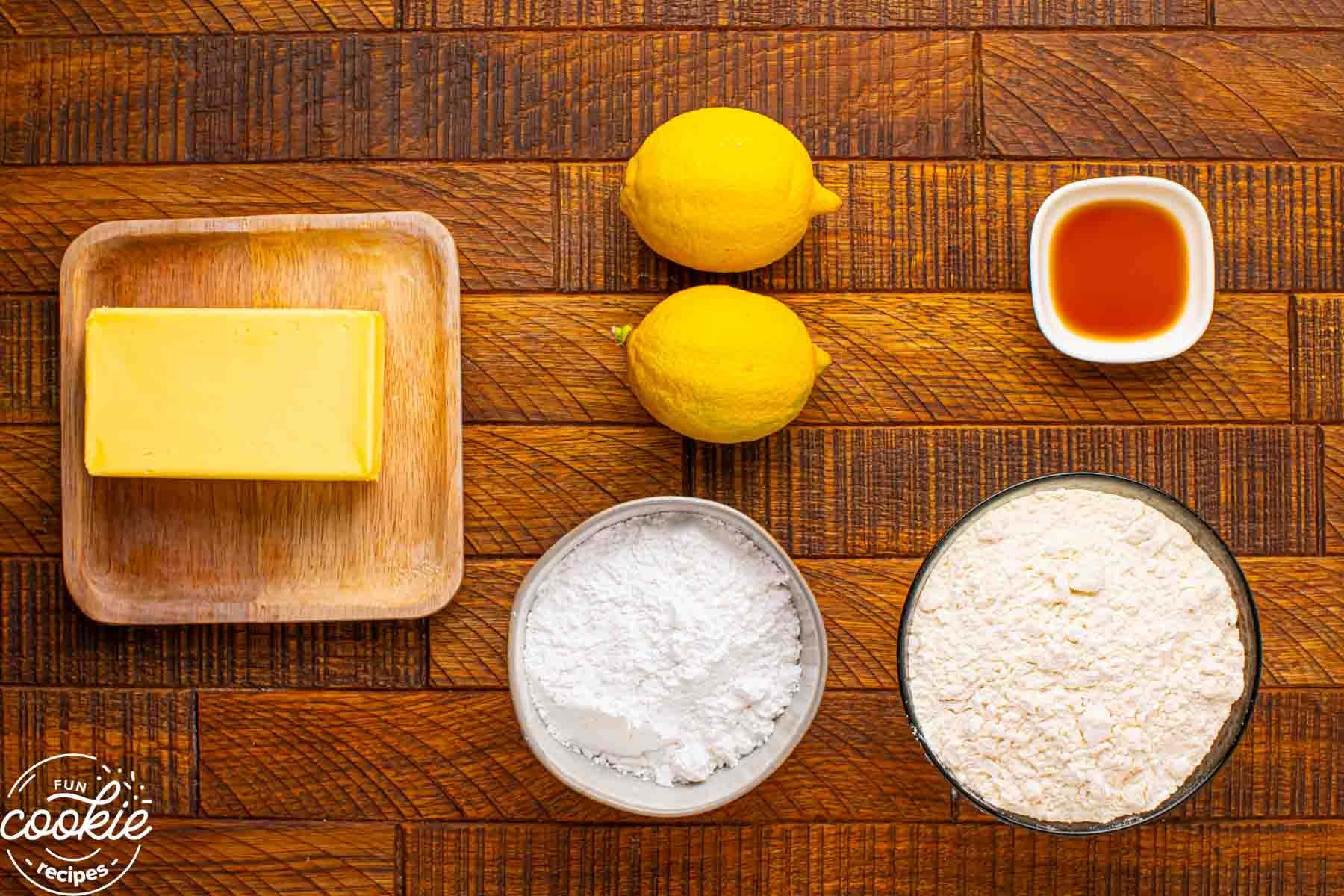 Overhead view of ingredients for lemon snowball cookies including butter, lemons, powdered sugar, flour, and vanilla extract on a wooden surface