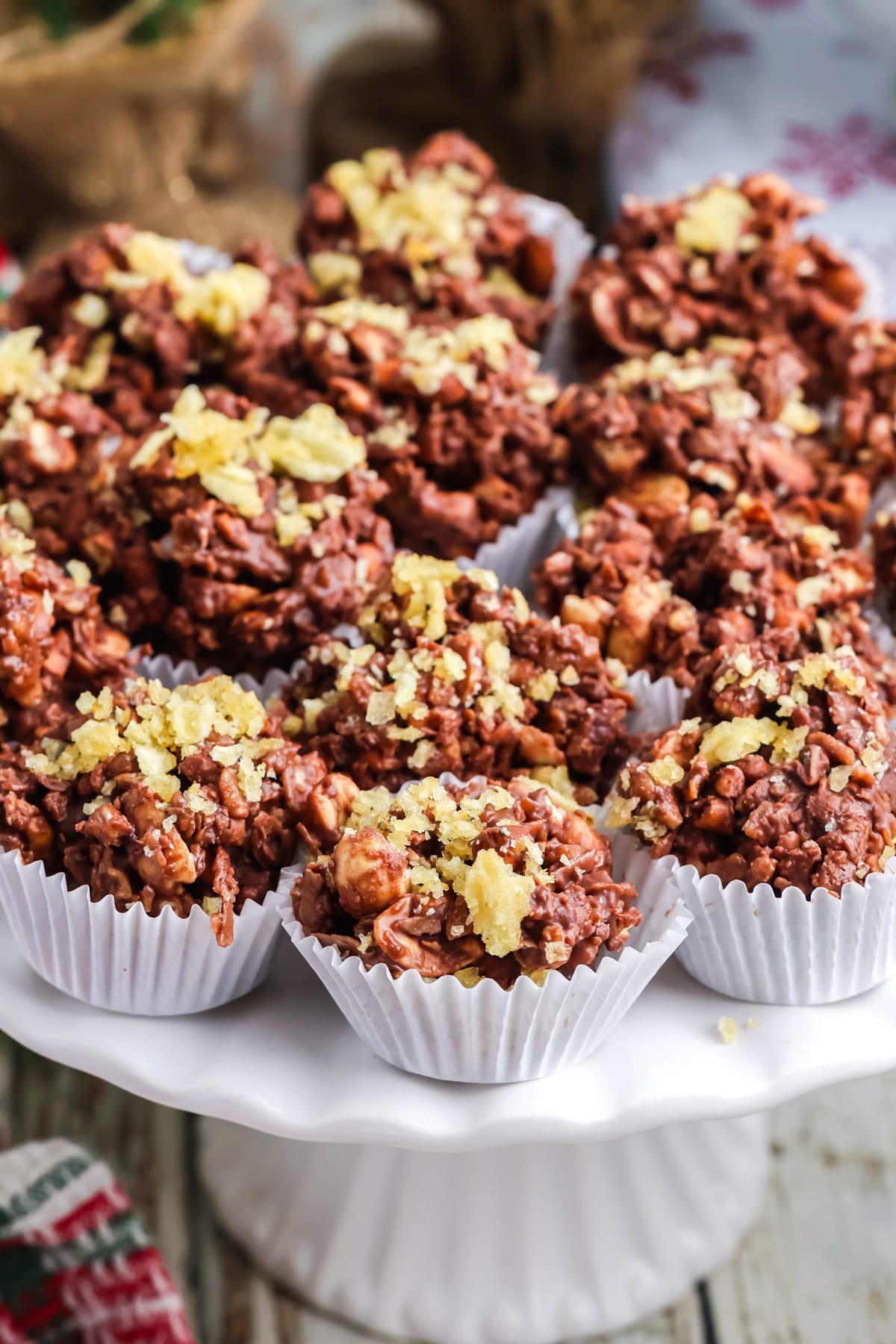 Chocolate potato chip clusters arranged on a white pedestal stand with festive holiday decorations in the background.